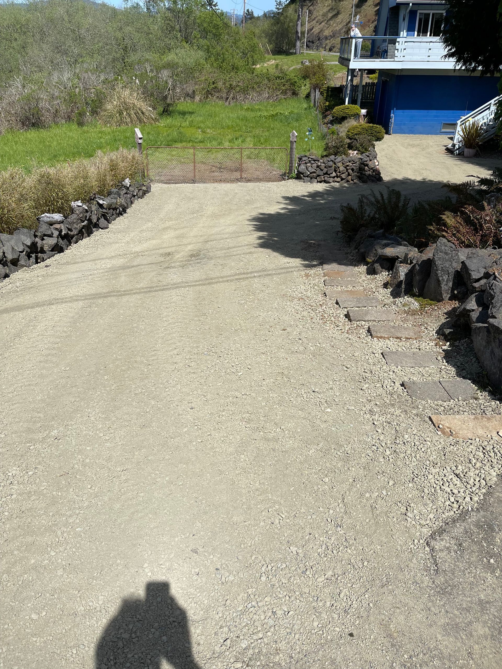 A shadow of a person is cast on a gravel driveway leading to a house.