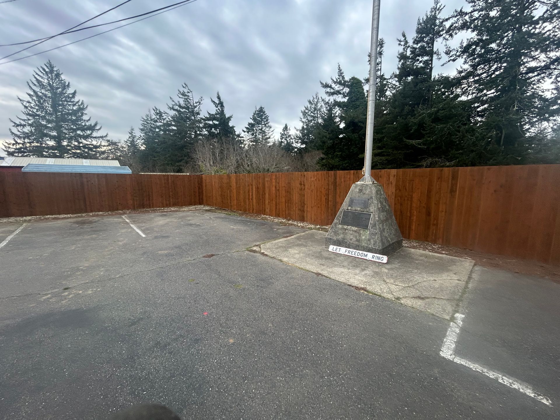 Empty parking lot with painted spaces, wooden fence, trees, and a basketball hoop pole on a cloudy day