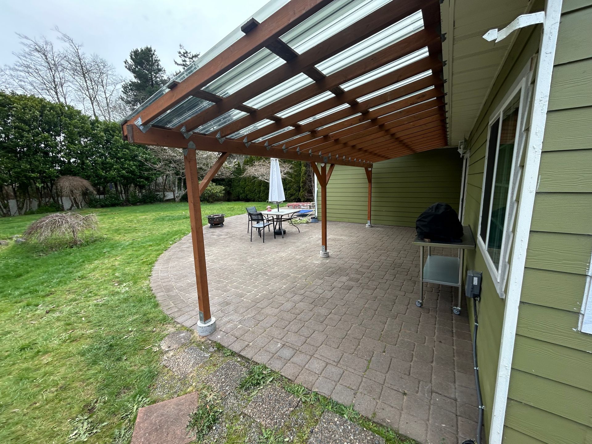 Covered patio with brick pavers, outdoor table and chairs, and a grassy yard beside a green house.
