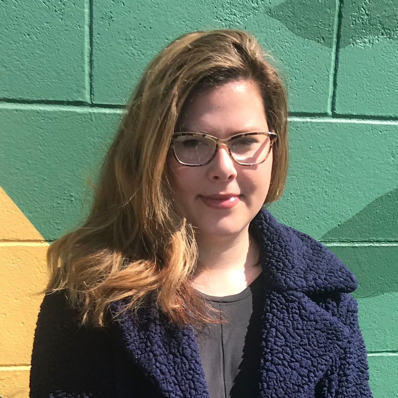 A woman wearing glasses and a blue coat is standing in front of a green wall.