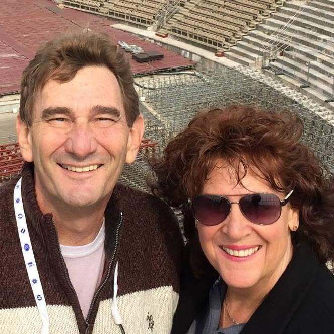 Smiling couple wearing sunglasses, posing for a selfie in an amphitheater seating area.
