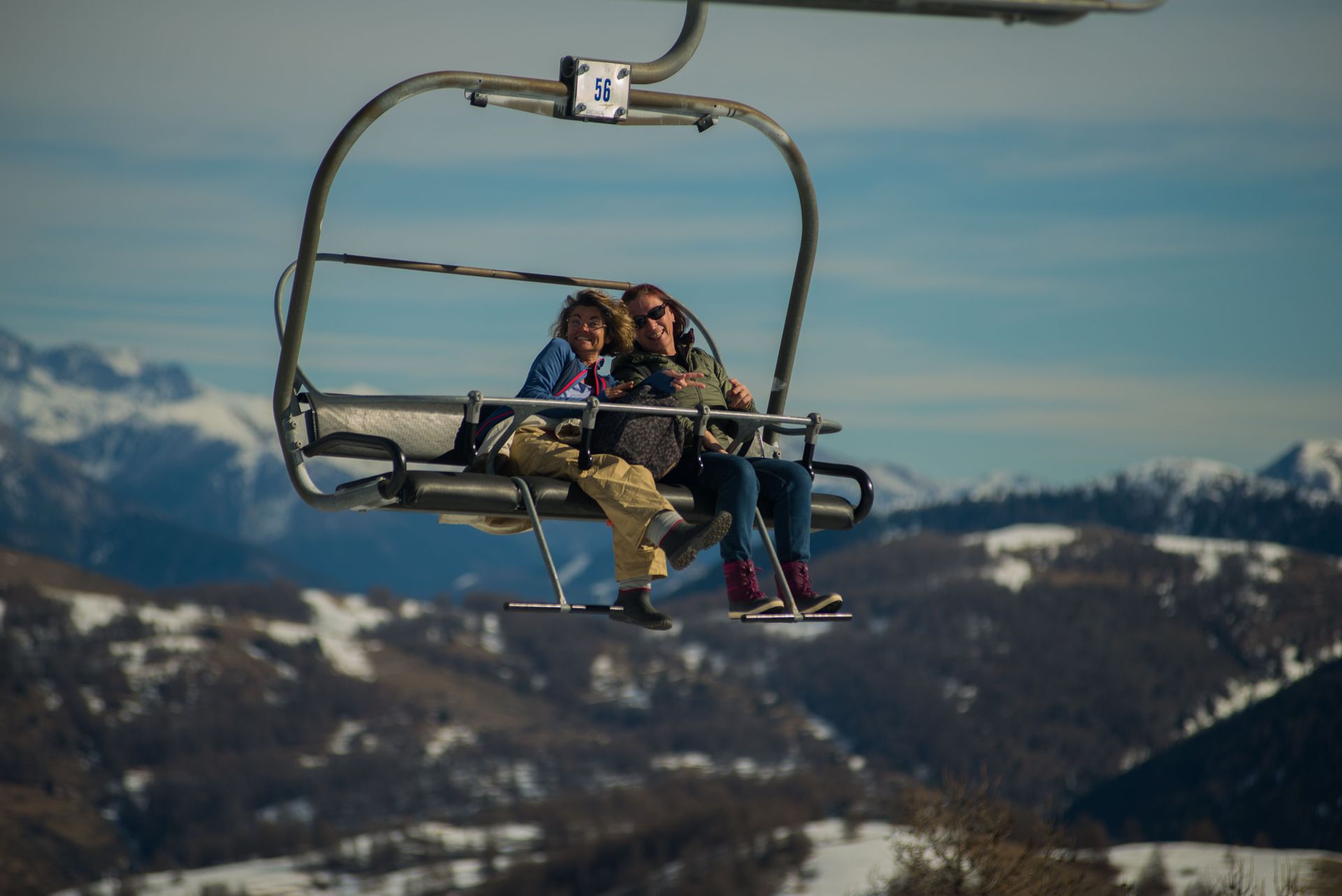 Two people riding a ski lift above snowy mountains under a blue sky