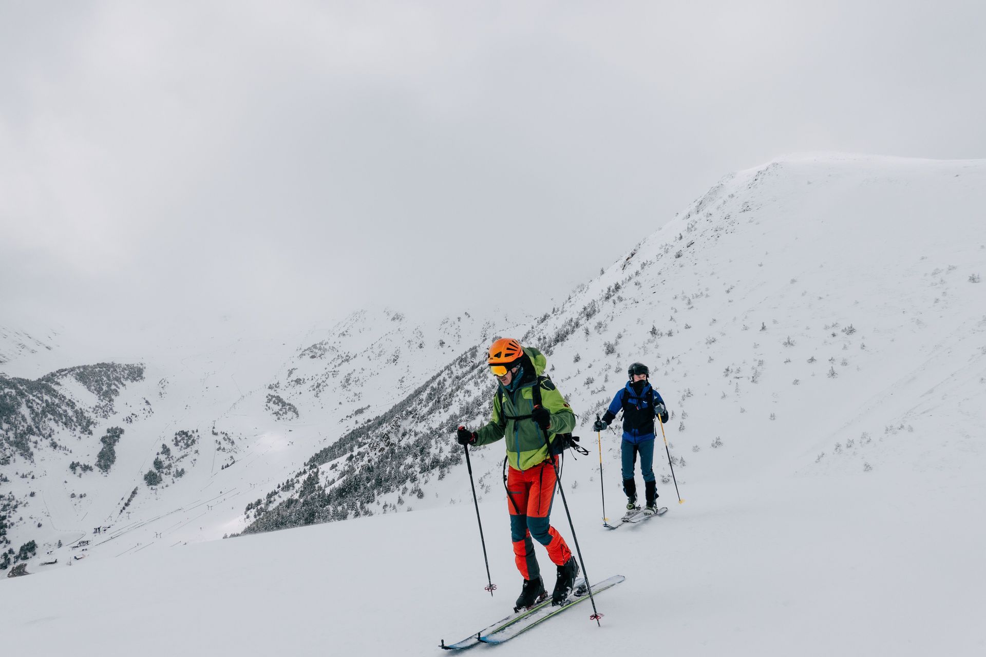Two skiers in colorful gear traverse a snowy mountain slope under an overcast sky