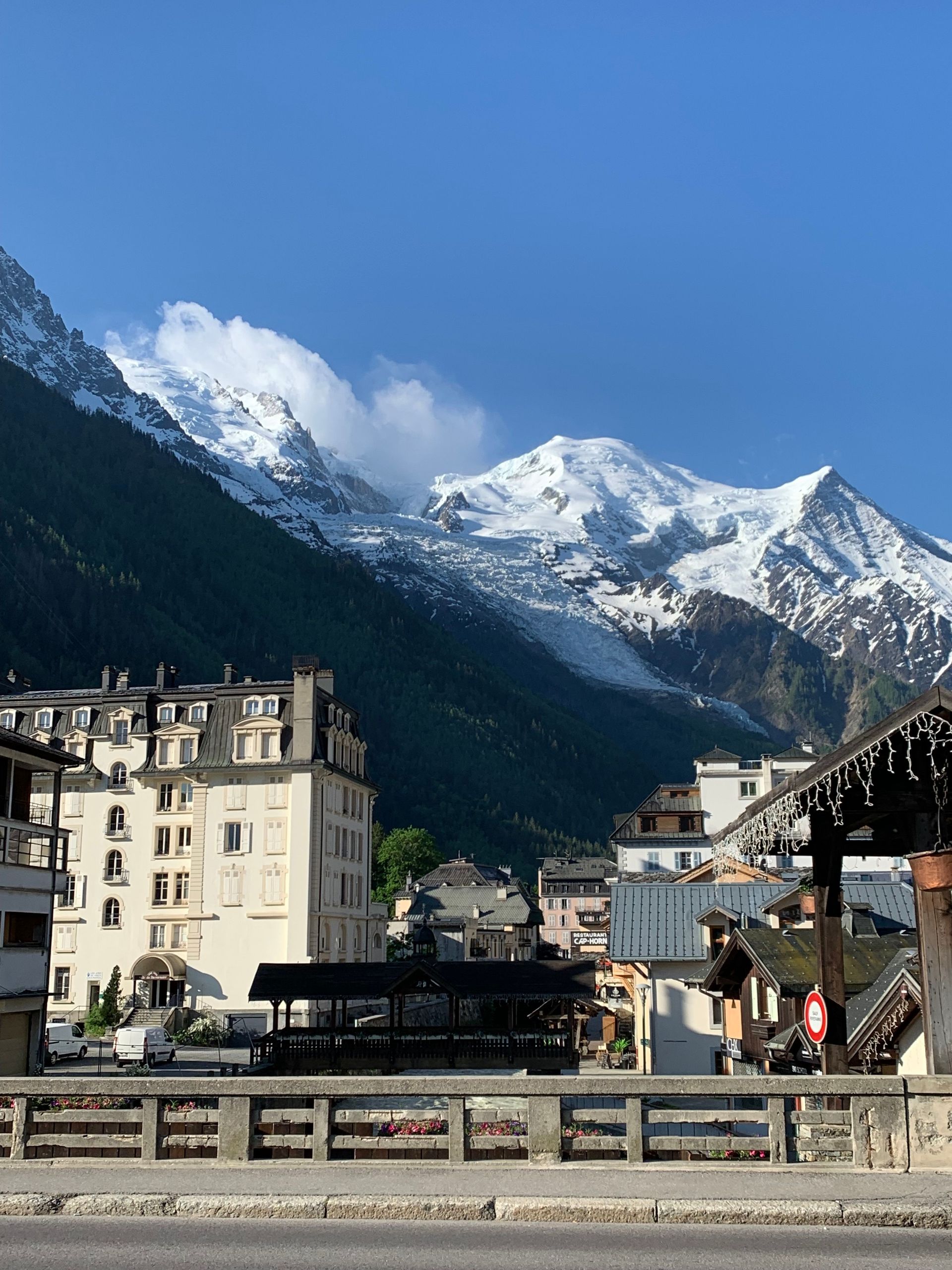 Snowy mountain peaks above a valley town with buildings and a bridge under a clear blue sky