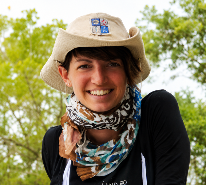 Smiling person in a tan hat and patterned scarf outdoors with green trees in the background