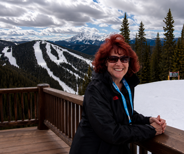 Smiling woman with red hair on a snowy mountain deck, ski slopes and pine trees behind her