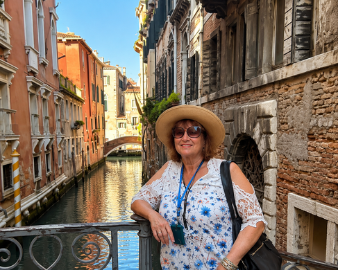 Woman in a straw hat and sunglasses standing on a bridge by a Venice canal, with colorful buildings behind her