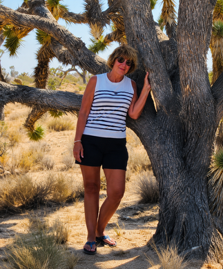 Woman in a striped tank top and black shorts leaning against a desert tree in sunglasses