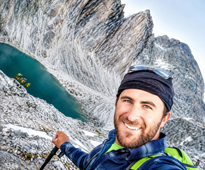 Smiling hiker taking a selfie beside a turquoise mountain lake and steep rocky cliffs