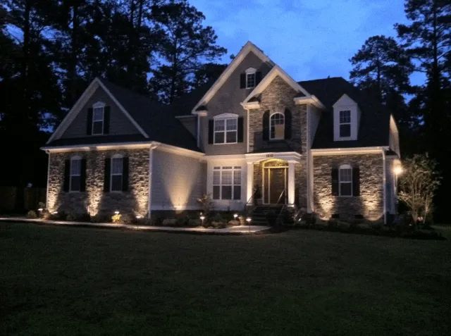 Two-story house with exterior lighting illuminating stone facade and landscaping at dusk.