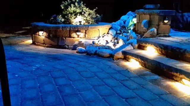 Night-lit outdoor patio with stone pavers and retaining wall. Blue and warm yellow lights illuminate the area.