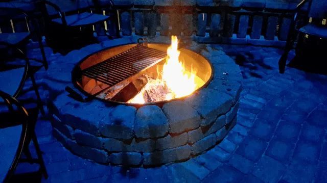 Fire pit with grill, flames burning brightly; on a blue-lit brick patio.