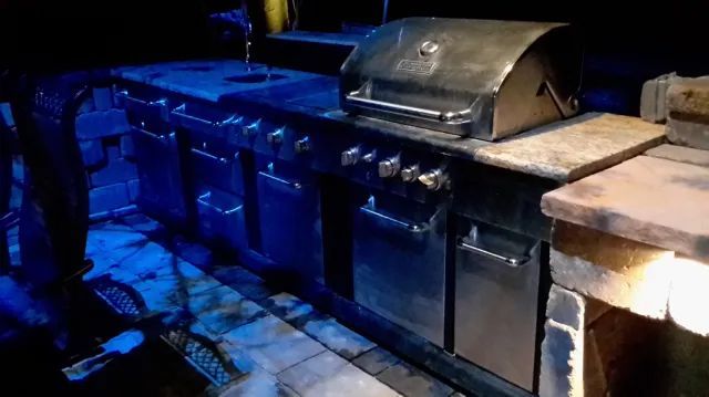 Outdoor kitchen with a large grill and built-in components, lit by blue lights, in a dark setting.