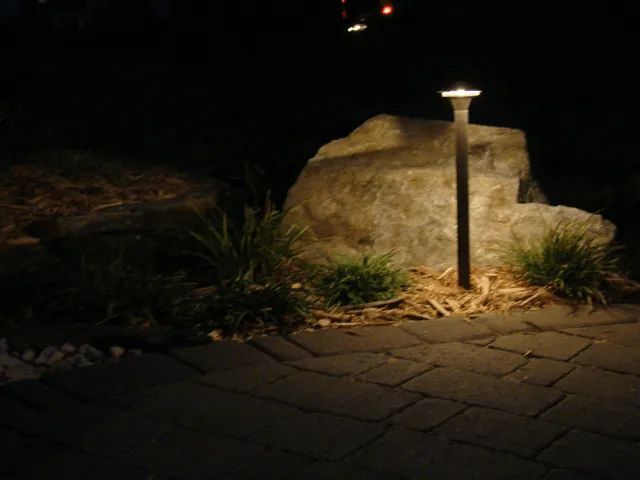 Nighttime landscaping illuminated by a small lamp; large rock, grass, and paved walkway.