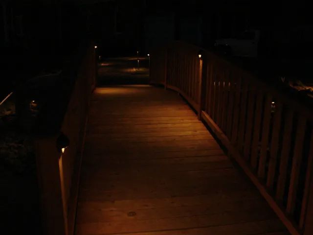 Wooden bridge at night, lit by small, warm lights along the path and railing, dark surroundings.