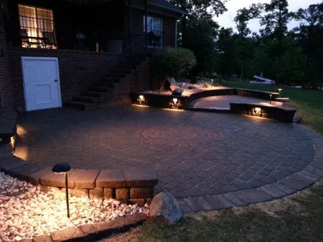 Nighttime shot of a brick patio with built-in seating, illuminated by low lights, next to a house.