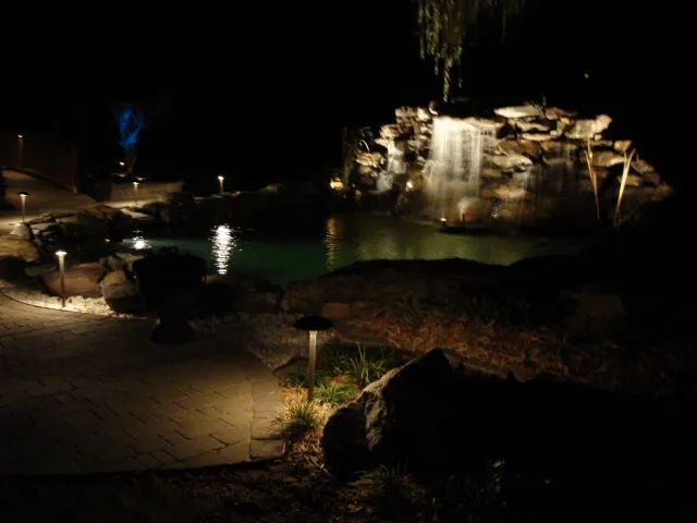 Nighttime pond and waterfall scene illuminated by landscape lighting.