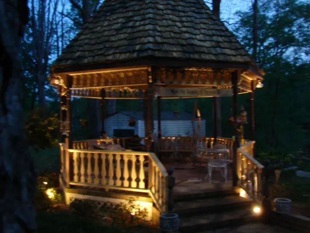 Lit-up gazebo with ornate details, steps, and surrounding trees at dusk.