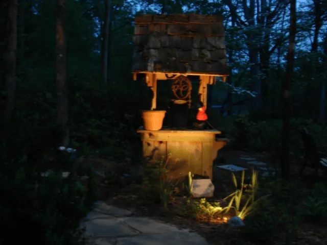 Stone wishing well illuminated at night in a wooded area; bucket and red lantern visible.