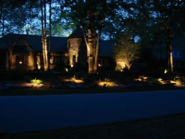 Nighttime view of a house illuminated by outdoor landscape lighting. Trees and bushes surround the home.