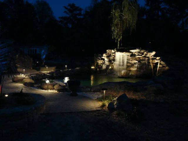 Nighttime view of a lit-up waterfall and pond in a garden with pathway lights illuminating the rocks.
