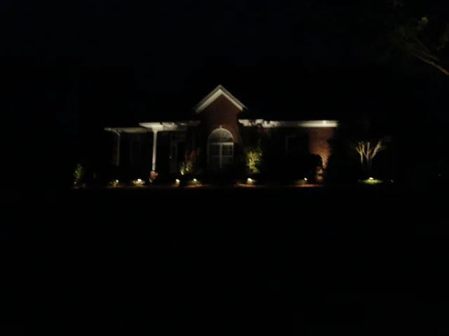 House at night, lit by landscape lights. Brick exterior, porch and small gabled roof.