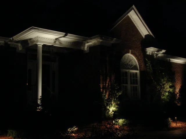 Night-lit brick house exterior. Upward lighting on the building facade, bushes, and porch columns.
