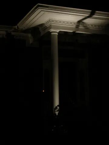 Architectural detail of a white column and ornate ceiling illuminated against a dark background.