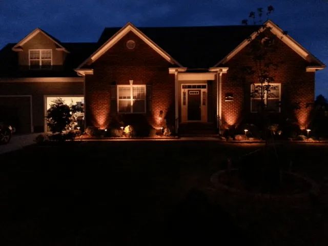 A brick house at night with landscape lighting, illuminating the facade and surrounding lawn.