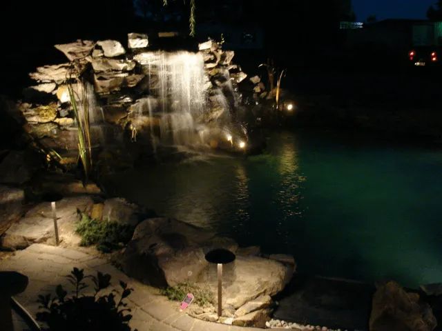 Waterfall at night, illuminated by soft lights. Water cascades into a dark pond surrounded by rocks and foliage.