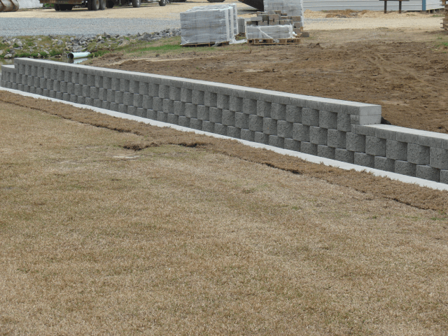 Gray retaining wall made of interlocking blocks on a grassy lawn. Construction site in the background.