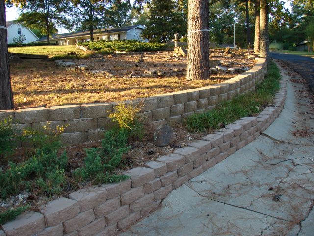 Brick retaining wall beside a paved driveway, with trees and a grassy area in the background.