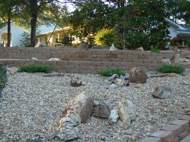 Stone terraced garden with gravel, rocks, and green shrubbery. Houses and trees in the background.