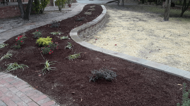 A winding garden bed with dark mulch and small plants, bordered by a stone wall and brick pathway.