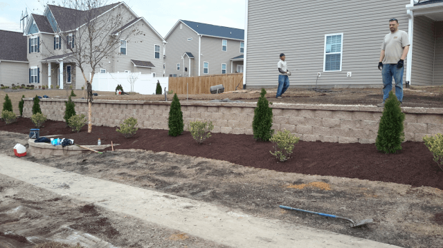 Two people beside a home with a decorative retaining wall and landscaping