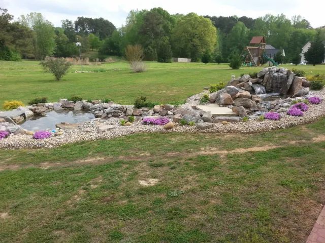 A backyard pond and waterfall feature surrounded by rocks, gravel, and purple flowers on a grassy lawn.