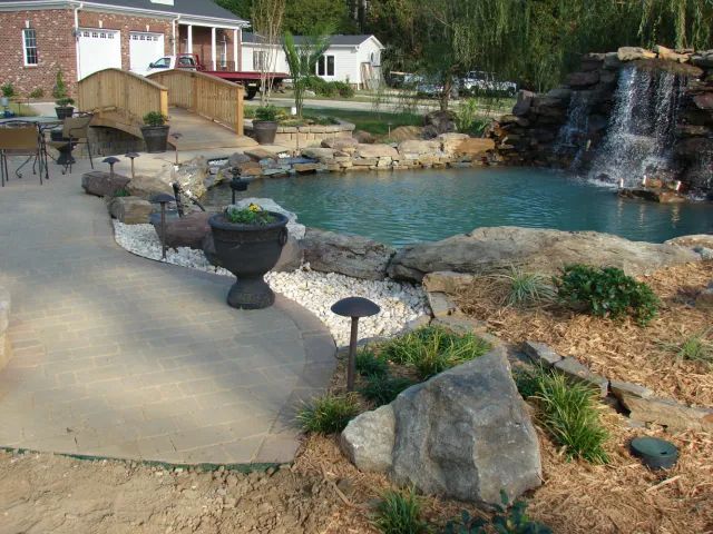 A backyard pond with a waterfall, bridge, and surrounding landscaping.