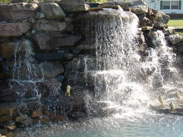 Waterfall cascading over dark rocks into a pool of water, sunny outdoor setting.