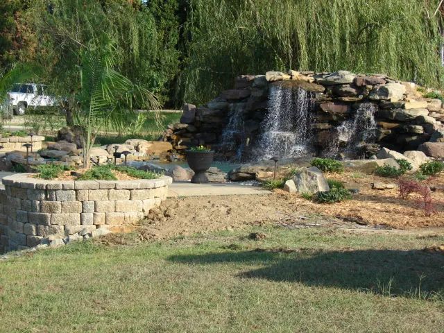 Waterfall feature in a landscaped yard, with a stone retaining wall and a car in the background.