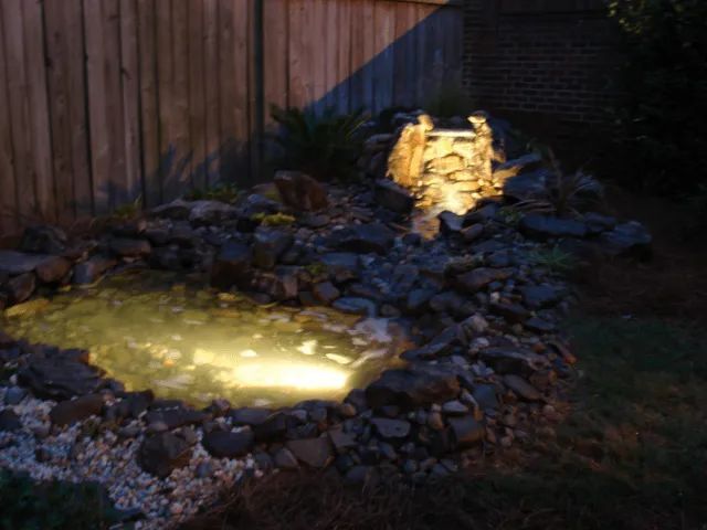 Lit-up backyard pond and waterfall feature at night, surrounded by rocks and a wooden fence.