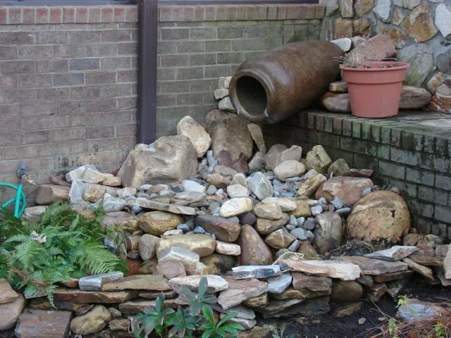 Water feature with rocks, plants, and a large ceramic pot spilling water. Brick and stone background.