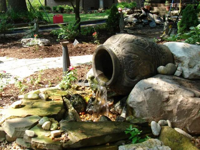 Water flows from a large, ornate urn into a stone-lined pond in a garden setting.