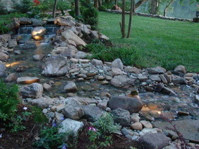 A rock-lined stream cascading down, lit with subtle lights, next to green grass.