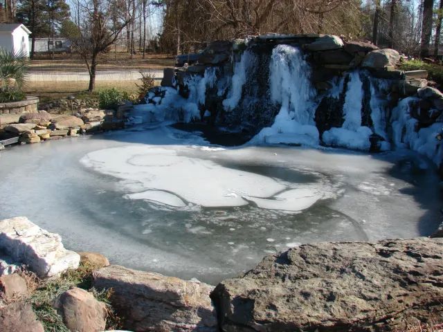 Frozen pond with a small waterfall, surrounded by rocks and a grassy bank.