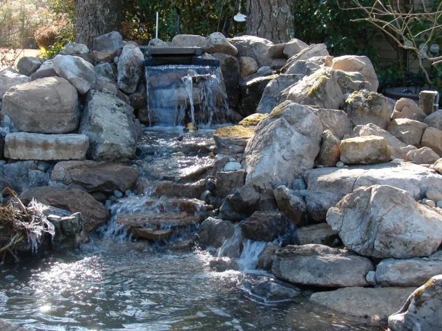 Rocky waterfall cascading into a pond, surrounded by large stones, sunlight glinting off the water.