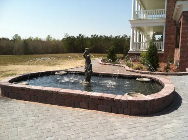 Fountain with stone border and statue in front of a brick house. Sprays of water, blue sky.