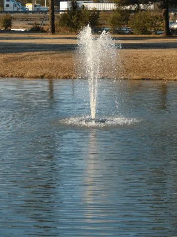 Fountain spraying water high into the air in a pond; brown bank and distant trees.