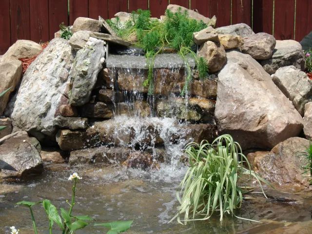 Small rock waterfall cascading into a pond, surrounded by stones and greenery.