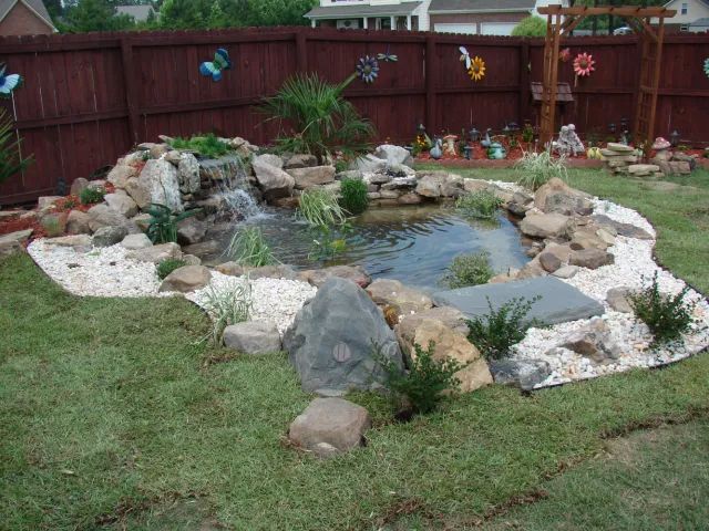Backyard pond with waterfall, surrounded by rocks and white gravel, lush green grass.