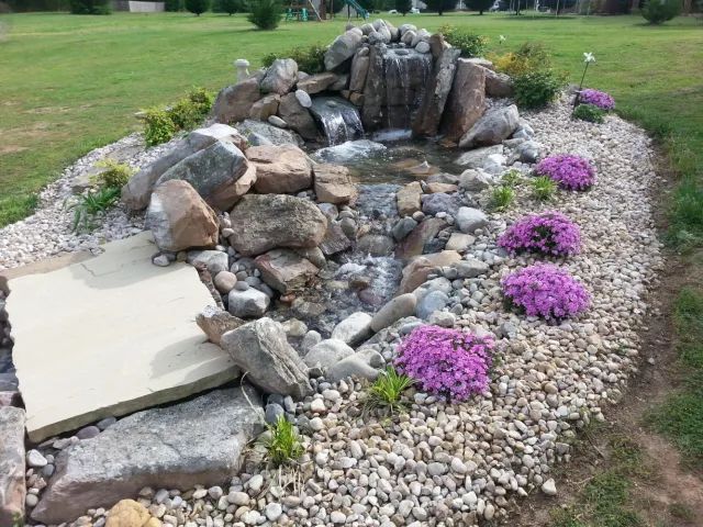 Water feature with waterfall, rocks, and purple flowers in a grassy yard.